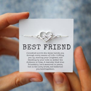 Op de afbeelding: Een zilverkleurige armband met een hartvormig oneindigheidssymbool. De armband is bevestigd aan een witte kaart met de tekst "BEST FRIEND". De kaart bevat ook een boodschap over vriendschap.