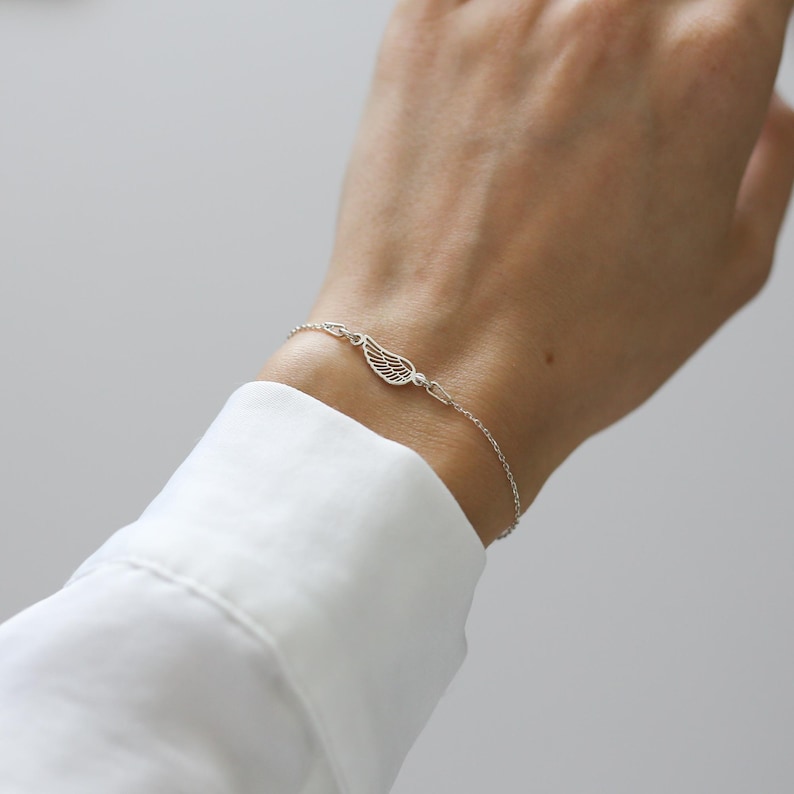 May include: A delicate silver bracelet featuring a wing charm and a small, round accent. The bracelet is worn on a wrist, showcasing its fine chain and intricate design. The wing charm is the focal point, adding a touch of elegance.