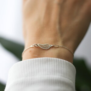 May include: A delicate silver bracelet featuring a wing charm. The wing is intricately detailed with fine lines, and the bracelet is fastened with a small clasp. The bracelet is worn on a wrist, against a white background.