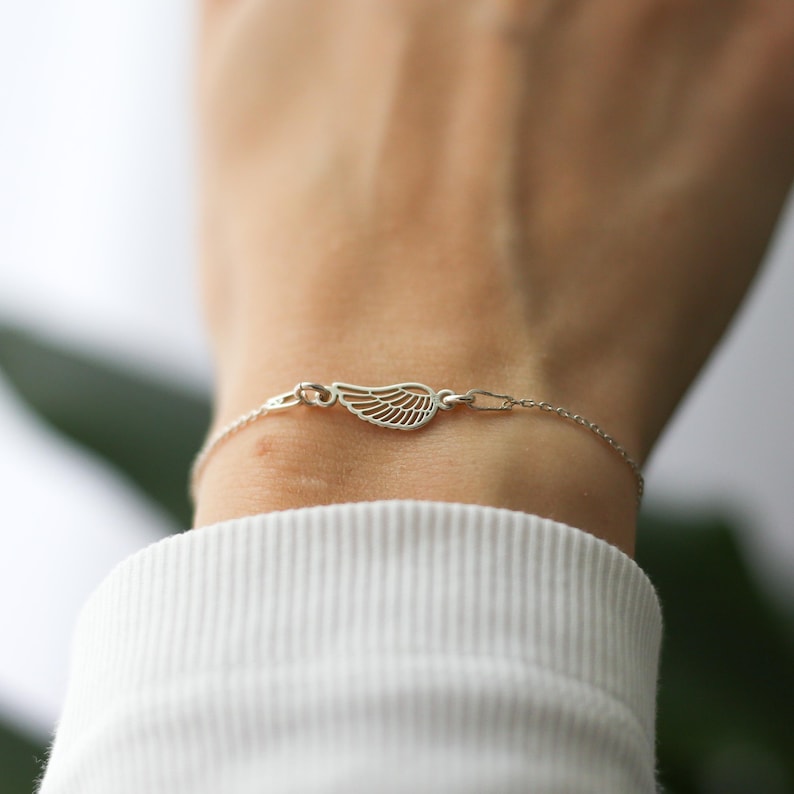 May include: A delicate silver bracelet featuring a wing charm. The wing is intricately detailed with fine lines, and the bracelet is fastened with a small clasp. The bracelet is worn on a wrist, against a white background.