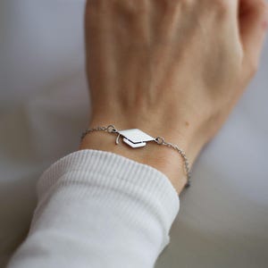 Puede incluir: Una pulsera plateada con un dije de birrete de graduación. El dije es una representación detallada en miniatura de un birrete. La pulsera se lleva en una muñeca, con una camisa blanca de manga larga en el fondo.