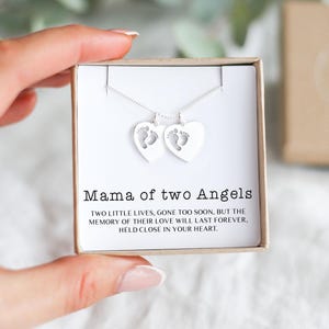 Op de afbeelding: Een zilveren ketting met twee hartvormige hangers, elk met een babyvoetafdruk. De ketting wordt gepresenteerd in een geschenkdoos met de tekst "Mama of two Angels".