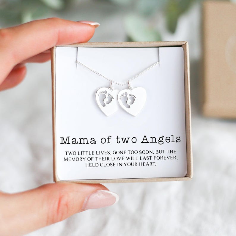 Op de afbeelding: Een zilveren ketting met twee hartvormige hangers, elk met een babyvoetafdruk. De ketting wordt gepresenteerd in een geschenkdoos met de tekst "Mama of two Angels".