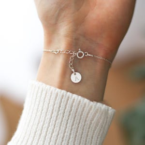 May include: A delicate silver bracelet with a small circular charm engraved with the letter "A". The bracelet is attached to the wrist with a clasp and chain. The person is wearing a white sweater.