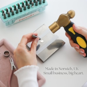 May include: A person using a hammer to stamp a small metal pendant. The image shows tools including a hammer, metal stamps, and a tray with jewelry components. Text in the image reads "Made in Norwich, UK - Small business, big heart."