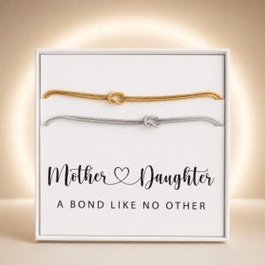 May include: Two thin chain bracelets, one gold-toned and one silver-toned, each featuring a central knot design. They are presented in a white box with the black printed text: Mother Daughter A BOND LIKE NO OTHER, featuring a heart symbol.
