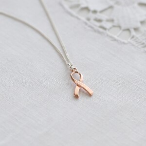 May include: A silver chain necklace featuring a rose gold ribbon pendant. The ribbon is a symbol of support and awareness. The necklace is presented on a white, textured background.