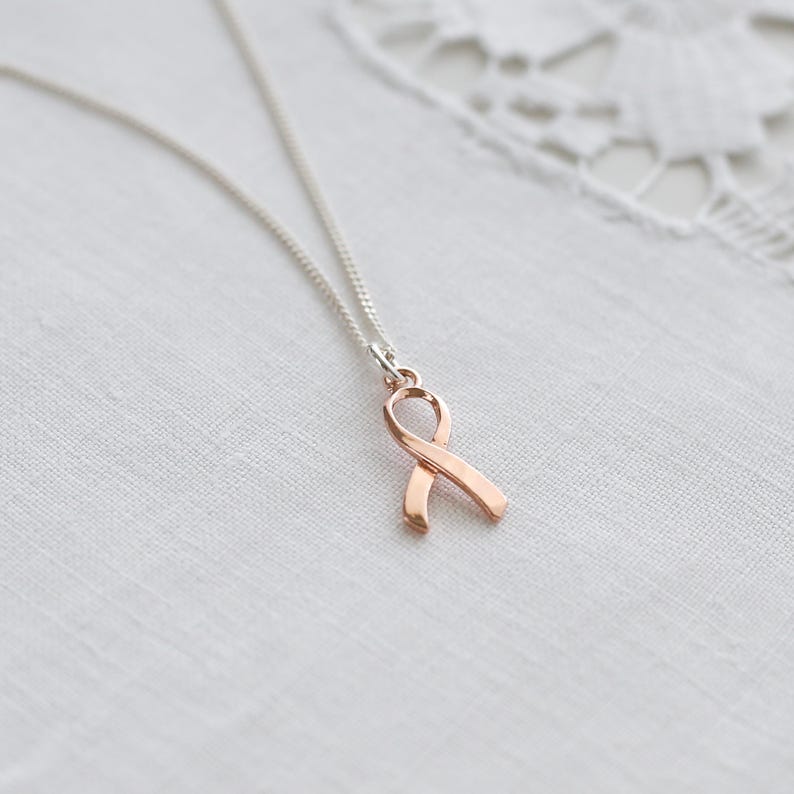 May include: A silver chain necklace featuring a rose gold ribbon pendant. The ribbon is a symbol of support and awareness. The necklace is presented on a white, textured background.