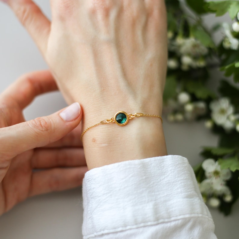 May include: A delicate gold bracelet featuring a round, emerald-green gemstone. The bracelet is worn on a wrist, with a white shirt visible in the background. The chain is fine and the gemstone is set in a gold bezel.