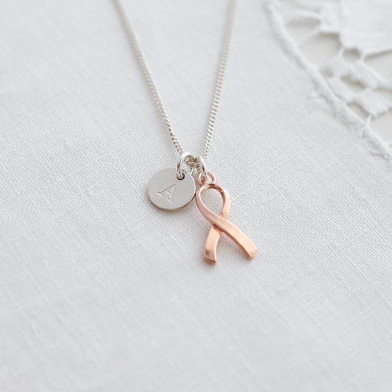 May include: A silver necklace featuring a small, circular pendant with the letter "A" and a rose gold ribbon charm. The ribbon is a symbol of awareness. The necklace is presented on a white background.