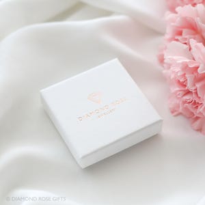 Op de afbeelding: Een witte sieradendoos met het logo "DIAMOND ROSE JEWELLERY" in ros&eacute;goud. De doos ligt op een witte satijnen ondergrond, met roze bloemen op de achtergrond. De doos is vierkant en heeft een getextureerd oppervlak.