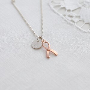 May include: A silver necklace featuring a small, circular pendant with the letter "A" and a rose gold ribbon charm. The necklace is presented on a white, textured background.