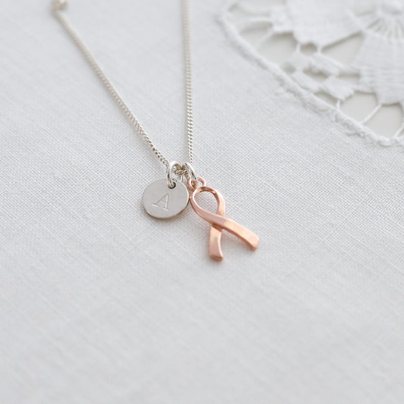 May include: A silver necklace featuring a small, circular pendant with the letter "A" and a rose gold ribbon charm. The necklace is presented on a white, textured background.