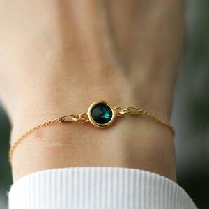 May include: A delicate gold bracelet featuring a round, dark teal gemstone set in a gold bezel. The bracelet has a fine chain and is secured with a small clasp. The gemstone is the focal point, adding a touch of color and elegance.