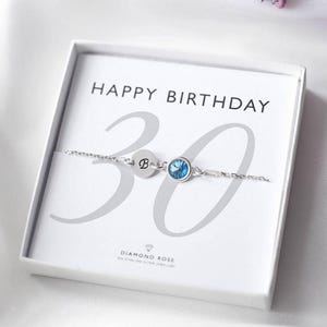 May include: A silver bracelet with a blue gemstone and the letter "B" is displayed inside a white gift box. The box reads "HAPPY BIRTHDAY" with a large "30" in the background. The bracelet is from Diamond Rose.