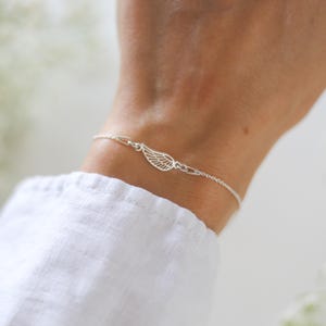 May include: A delicate silver bracelet featuring a small wing charm. The bracelet is made of a fine chain with a wing-shaped pendant. The bracelet is worn on a wrist, and the background is blurred.