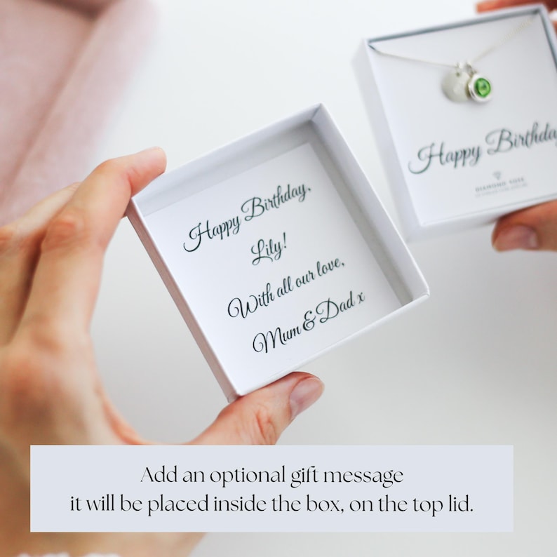 Op de afbeelding: Twee witte geschenkdozen worden opengehouden en onthullen een ketting en een gepersonaliseerde boodschap. De boodschap luidt "Gelukkige verjaardag, Lily! Met al onze liefde, Mama & Papa x." De ketting heeft een zilveren ketting en een groene edelsteen.