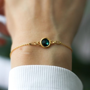 May include: A delicate gold bracelet featuring a round, dark green gemstone set in a gold bezel. The bracelet has a fine chain with small links and is worn on a wrist, against a white shirt.