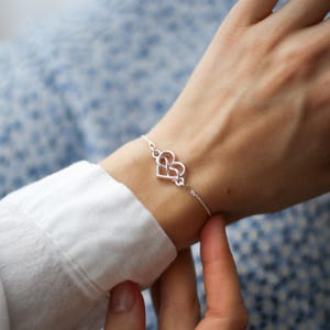 Puede incluir: Una delicada pulsera de tono plateado que presenta un dije central en forma de coraz&oacute;n con un intrincado dise&ntilde;o de nudo infinito. El dije est&aacute; conectado a una cadena fina y pulida que rodea la mu&ntilde;eca, creando una apariencia simple y refinada.