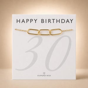 Op de afbeelding: Een goudkleurige kettingarmband met drie langwerpige ovale schakels, gepresenteerd op een witte kaart met de tekst HAPPY BIRTHDAY, een groot grijs getal 30 en de merknaam DIAMOND ROSE in het zwart onderaan de kaart gedrukt.