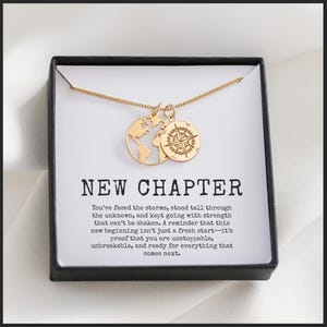 May include: Gold-colored necklace with two pendants: a globe and a compass. The necklace is displayed in a black box with the words "NEW CHAPTER" and inspirational text. The box is on a white surface.