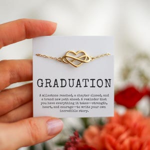 May include: A gold-colored bracelet with a heart-shaped infinity symbol is displayed on a white card. The card reads "GRADUATION" and includes a motivational message. The background features blurred flowers.