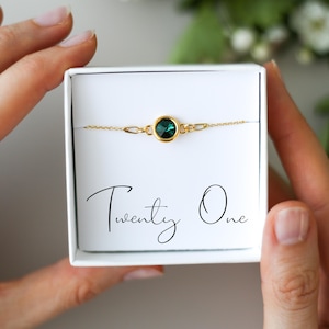 Op de afbeelding: Een goudkleurige armband met een ronde, smaragdgroene edelsteen wordt gepresenteerd in een witte geschenkdoos. De doos heeft de tekst "Twenty One" in cursief. De armband wordt door een persoon vastgehouden.