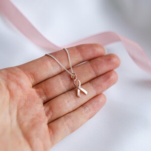 May include: A silver chain necklace featuring a rose gold ribbon pendant. The ribbon is a symbol of awareness, set against a soft, blurred background with a pink ribbon. The necklace is displayed on a hand.