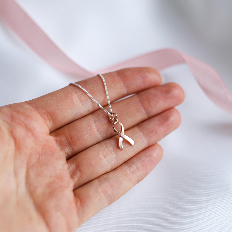 May include: A silver chain necklace featuring a rose gold ribbon pendant. The ribbon is a symbol of awareness, set against a soft, blurred background with a pink ribbon. The necklace is displayed on a hand.