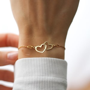 May include: A gold bracelet with two intertwined heart-shaped pendants. The fine chain links the hearts, forming a simple, elegant design. The bracelet is worn on a wrist, against a white long-sleeved top.