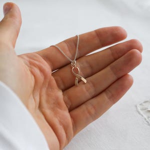 May include: A silver chain necklace with a rose gold ribbon pendant. The ribbon is a symbol of awareness. The necklace is displayed on a person's open palm against a white background. The pendant is approximately 2 cm in length.