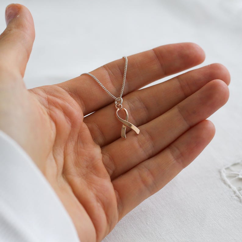 May include: A silver chain necklace with a rose gold ribbon pendant. The ribbon is a symbol of awareness. The necklace is displayed on a person's open palm against a white background. The pendant is approximately 2 cm in length.