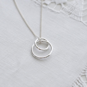 May include: A silver necklace with two interlinked rings. The pendant hangs from a fine chain, offering a minimalist and elegant aesthetic. The rings have a hammered texture, adding visual interest to the jewellery.