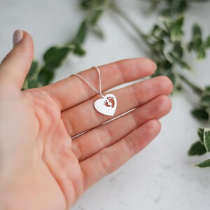 Op de afbeelding: Een zilveren hartvormige hangerketting met kleine voetstappen in het hart. De ketting wordt in een hand gehouden, met groene bladeren op de achtergrond. De hanger is ongeveer 2 cm breed.