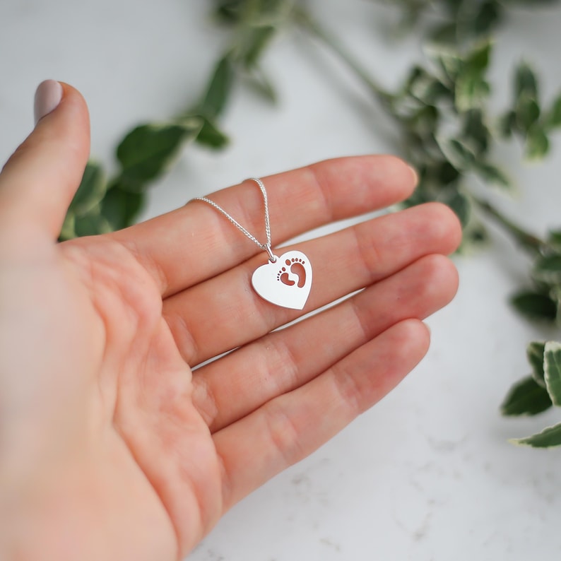 Op de afbeelding: Een zilveren hartvormige hangerketting met kleine voetstappen in het hart. De ketting wordt in een hand gehouden, met groene bladeren op de achtergrond. De hanger is ongeveer 2 cm breed.