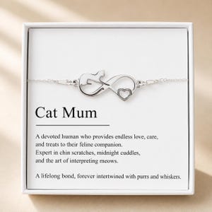May include: A silver-toned bracelet featuring an infinity symbol charm integrated with a cat silhouette and a small heart. The jewelry is presented in a white box with the text Cat Mum and a definition printed in black ink on the inner card.