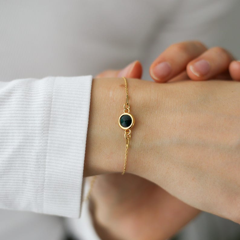 May include: A delicate gold bracelet featuring a dark green gemstone. The bracelet is a simple chain design with a single, round gemstone pendant. The jewelry is worn on a wrist, with a white shirt sleeve visible in the background.