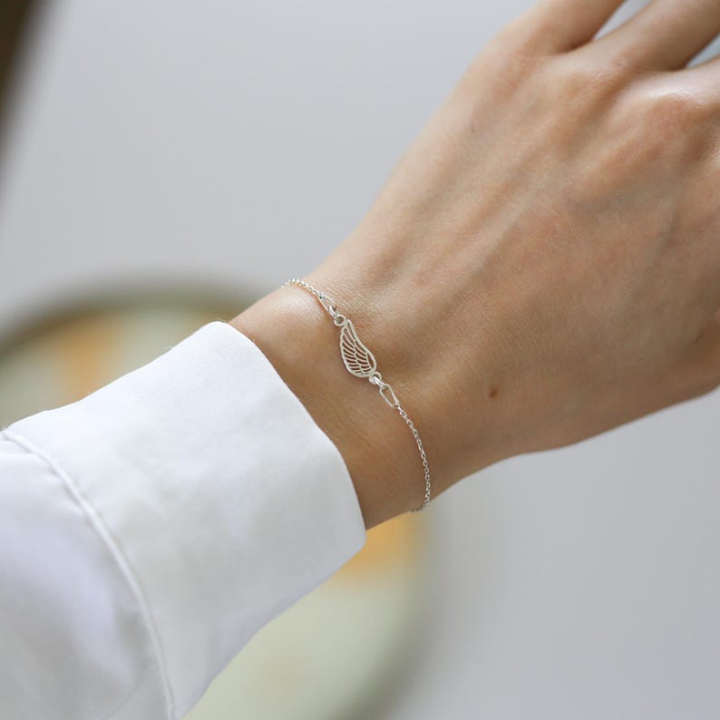 May include: A delicate silver bracelet featuring a small, openwork wing charm. The bracelet is worn on a wrist, with a white shirt sleeve in the background. The wing charm is the focal point, showcasing intricate details.