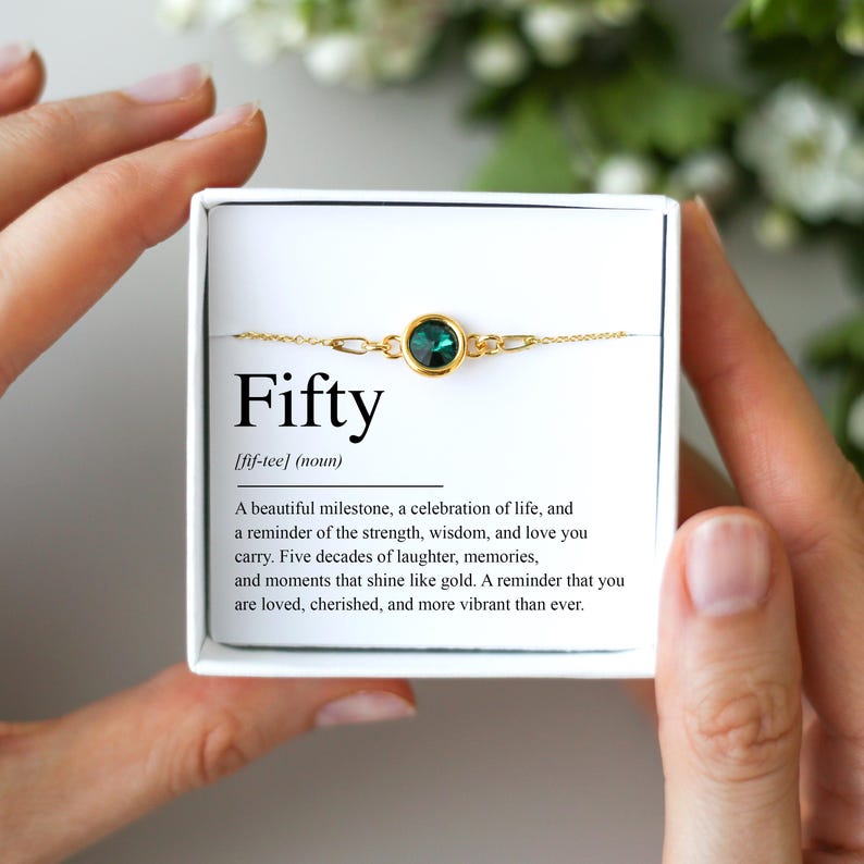 May include: A gold-toned bracelet with a round, teal-colored gemstone is displayed in a white box. The box has the word "Fifty" printed on it, along with a definition and a short message. The bracelet is a delicate piece of jewelry.