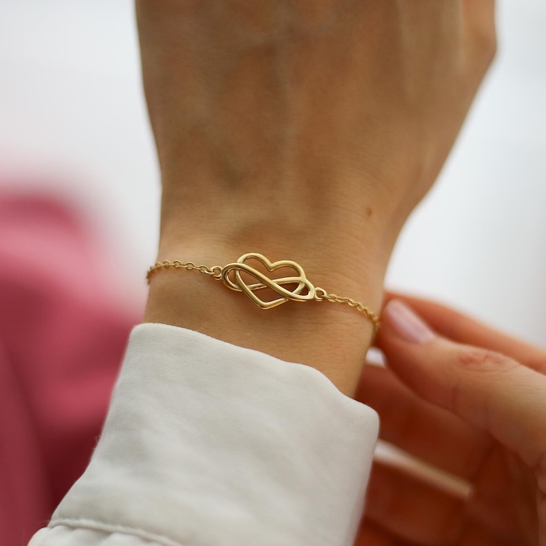 May include: A delicate gold-colored bracelet featuring an infinity symbol intertwined with a heart shape. The bracelet is on a slender chain and is being worn on a wrist. The jewelry is a romantic accessory.