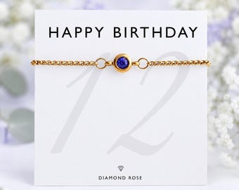 Op de afbeelding: Een goudkleurige kettingarmband met een enkele ronde blauwe kristallen steen in het midden, gepresenteerd op een witte kaart met de tekst HAPPY BIRTHDAY, een groot getal 12 en de merknaam DIAMOND ROSE met een klein diamanticoon onderaan.