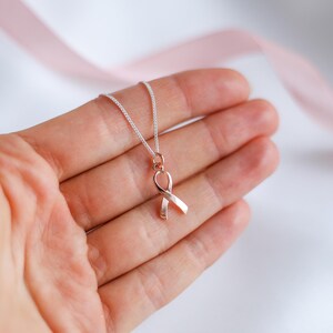 May include: A silver chain necklace with a rose gold ribbon pendant. The ribbon is a symbol of awareness. The necklace is displayed on a light background with a pink ribbon in the background.