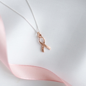 May include: A rose gold ribbon pendant necklace on a silver chain. The ribbon is a symbol of awareness, set against a white background with a pink ribbon in the foreground. The necklace is a delicate piece of jewellery.
