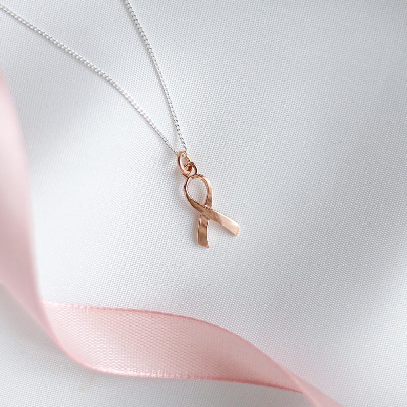 May include: A rose gold ribbon pendant necklace on a silver chain. The ribbon is a symbol of awareness, set against a white background with a pink ribbon in the foreground. The necklace is a delicate piece of jewellery.
