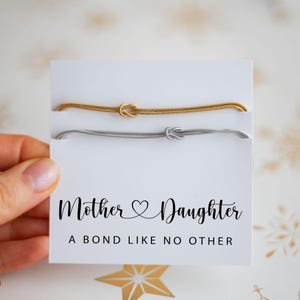 May include: A gold and silver bracelet set with a knot design, presented on a white card. The card features the text "Mother & Daughter, A Bond Like No Other" in black script. The bracelets are held by a hand.