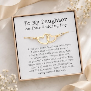 Puede incluir: Una pulsera de tono dorado con dos dijes de corazones entrelazados, presentada en una caja de regalo cuadrada. La tarjeta interior dice: To My Daughter on Your Wedding Day, seguida de un mensaje sentimental sobre el amor y el futuro.