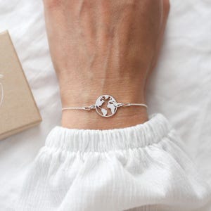 Può includere: Un braccialetto d'argento con un ciondolo a forma di globo. Il ciondolo rotondo presenta un design traforato della mappa del mondo. Il braccialetto è su un polso con una camicia bianca a maniche lunghe. Una piccola scatola marrone è sullo sfondo.