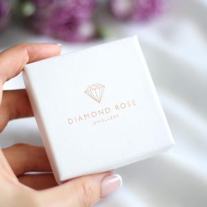 Op de afbeelding: Een witte sieradendoos met de woorden "DIAMOND ROSE JEWELLERY" in ros&eacute;goud gedrukt. De doos heeft een ros&eacute;gouden diamantafbeelding. De doos wordt in een hand gehouden.