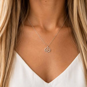 May include: A silver-coloured necklace featuring a delicate chain and a pendant shaped like an infinity symbol intertwined with a heart outline. The metallic pendant hangs centrally, showcasing a polished finish that reflects light against the white top.