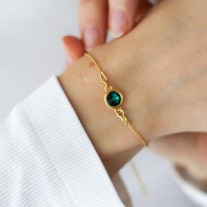 May include: Gold-toned bracelet featuring a round, emerald-green gemstone. The delicate chain is attached to a bezel setting that holds the gemstone. The bracelet is worn on a wrist, with a white shirt in the background.